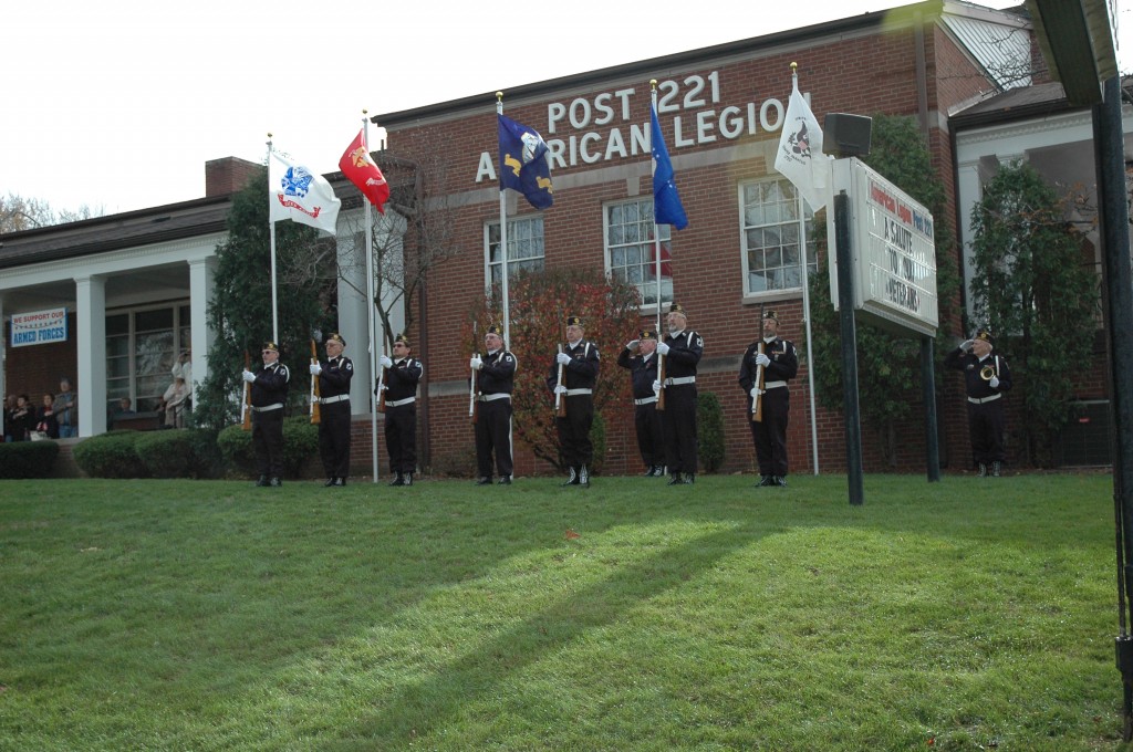 In front of the red-brick post building, seven members of the Color Guard stand in front of the legion's new five service flags.